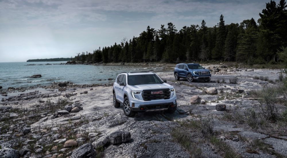 A white 2024 GMC Acadia AT4 and a blue 2024 GMC Acadia Denali parked on a beach.