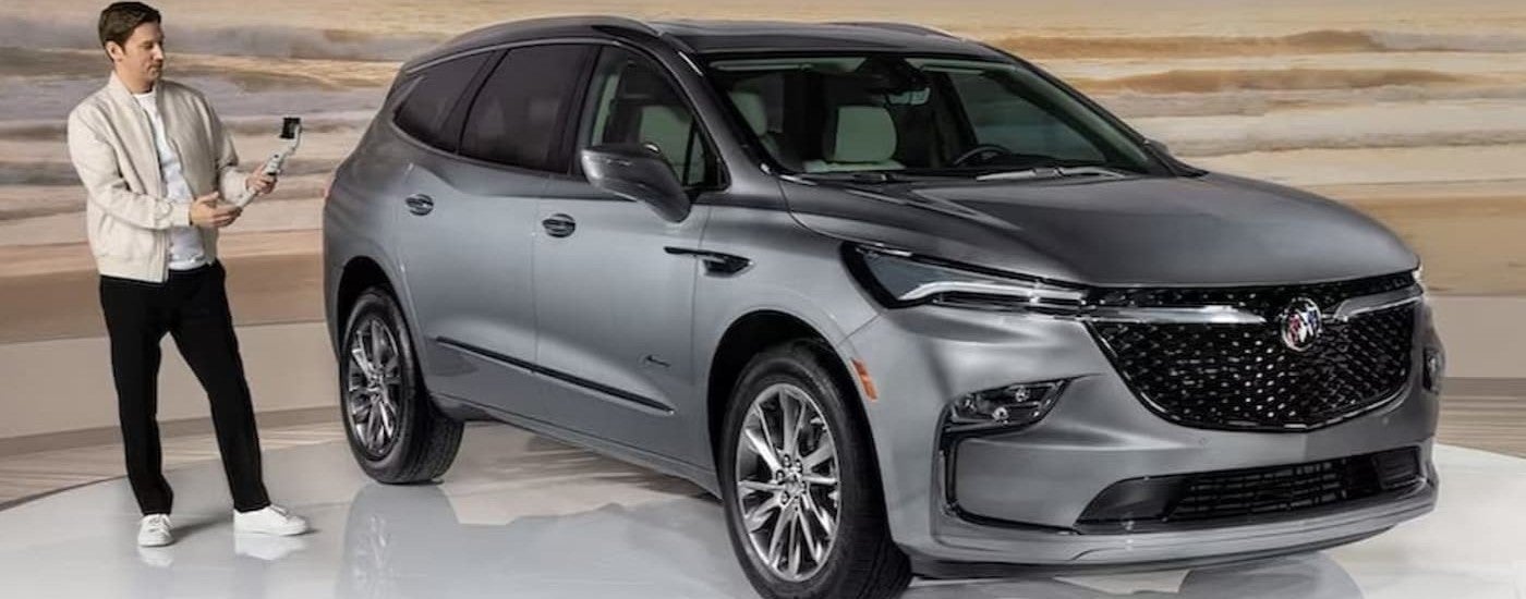 A person is shown standing next to a grey 2023 Buick Enclave parked in a showroom.