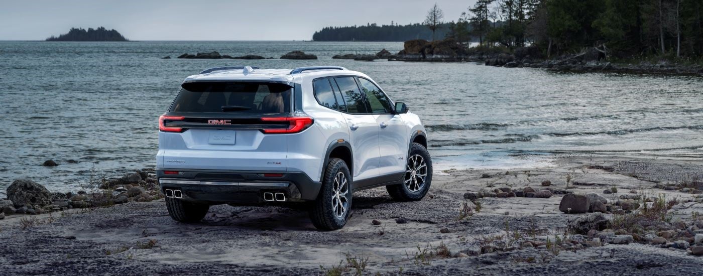 Rear angle view of a white 2024 GMC Acadia AT4 parked on a lake shore.